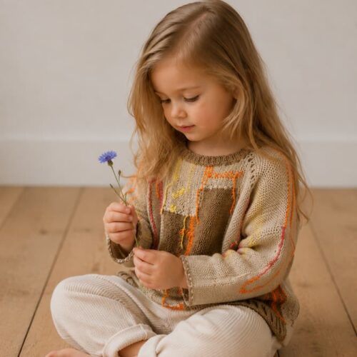 Handknitted baby sweater in natural tones with colorful accents, worn by a child sitting on a wooden floor