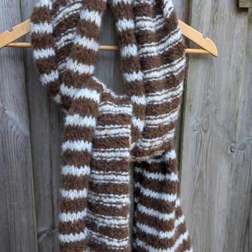Handknitted brown alpaca scarf by INESKA — hanging on a wooden hanger against a weathered fence